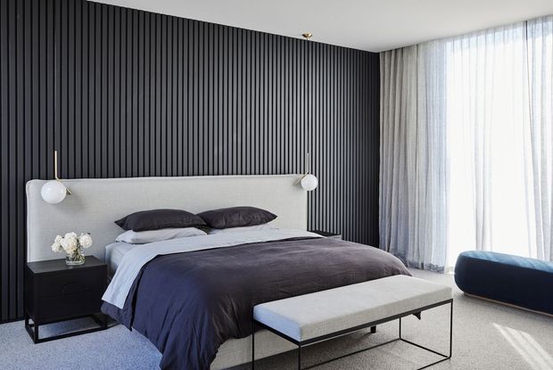 Contemporary bedroom with a dark vertically slatted feature wall, upholstered light-grey bedhead, dark navy and charcoal bedding, matching black bedside table, and a slim upholstered bench at the foot of the bed. Full-height sheer curtains provide soft daylight, creating a refined, high-contrast interior.