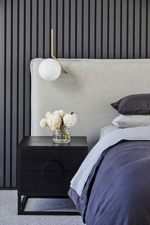 Close-up of a refined contemporary bedroom featuring a black vertical fluted feature wall, upholstered light-grey headboard, dark built-in-style bedside table, brass-and-white globe wall light, and layered bedding in soft grey, white, and deep charcoal-purple tones.