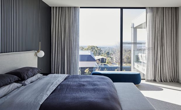 Contemporary bedroom with a large upholstered bed dressed in layered blue-grey bedding, a dark vertical slatted feature wall, full-height grey curtains, and expansive floor-to-ceiling glazing. A curved blue bench sits by the window, with a bright residential outlook beyond.