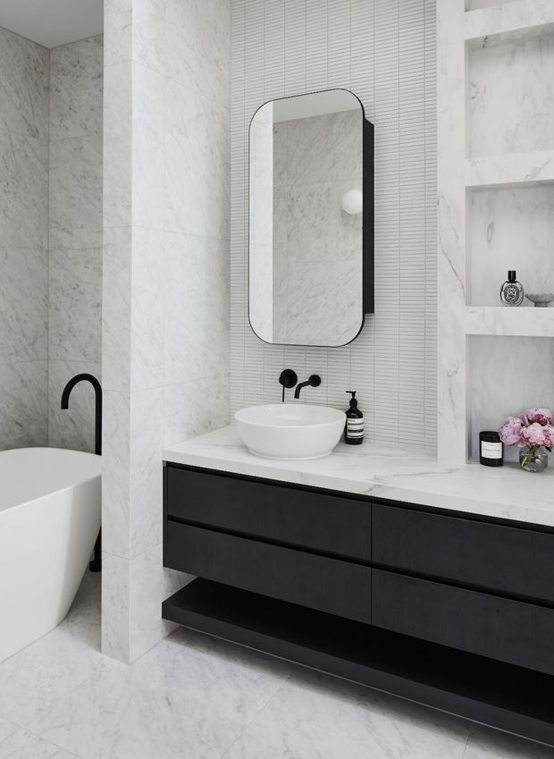 Contemporary bathroom with white marble-look wall and floor surfaces, a freestanding bathtub, black curved tapware, a wall-mounted black vanity with white benchtop and vessel basin, and a rounded rectangular mirror. A vertically ribbed tile splashback and recessed marble-look shelving add texture and storage.