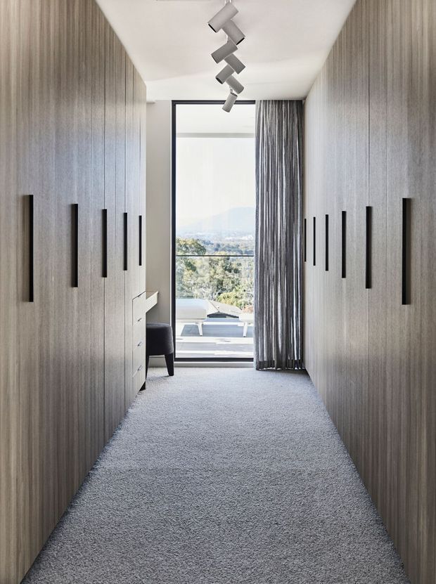 A narrow, refined dressing corridor or walk-in wardrobe lined on both sides with full-height grey wood-grain built-in cabinetry and long black handles. The space has a soft grey carpet, white ceiling, minimalist track lighting, and a floor-to-ceiling glazed opening with curtains at the end, overlooking a leafy landscape. A small white chest and upholstered chair are partially visible near the opening.