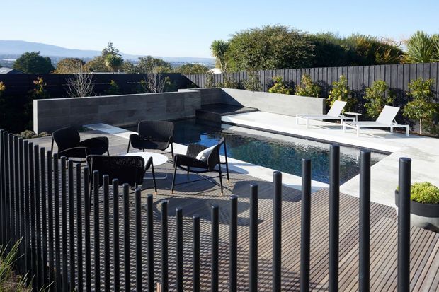 A contemporary landscaped outdoor pool area with a narrow rectangular swimming pool, pale concrete paving, dark rendered retaining walls, and a timber deck. Black vertical rod screening forms a sculptural foreground balustrade, while outdoor dining furniture and white sun loungers surround the pool. Dense planting and a distant hilly landscape are visible beyond the boundary.