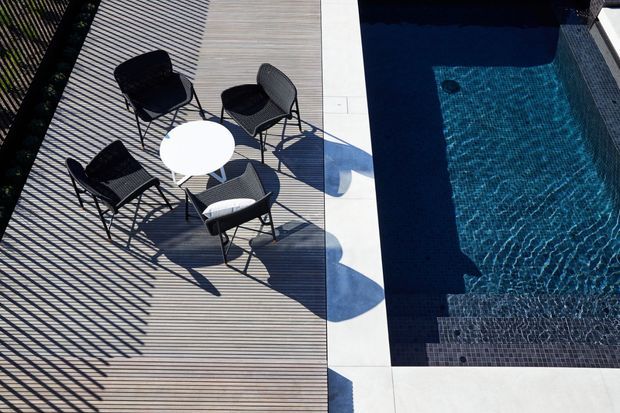 Overhead view of a contemporary poolside terrace with a rectangular swimming pool edged in pale paving. A small round white table and four dark woven outdoor chairs sit on a linear timber-look deck, with strong architectural shadows creating a graphic pattern across the space.
