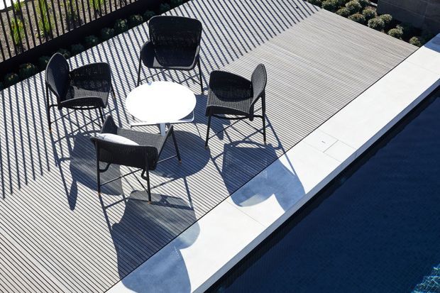 Elevated view of a contemporary outdoor terrace beside a dark swimming pool. The terrace is finished with narrow linear decking, bordered by pale rectangular coping or paving, and furnished with four black woven chairs around a small round white table. Strong diagonal shadows from a railing create a graphic pattern across the deck.