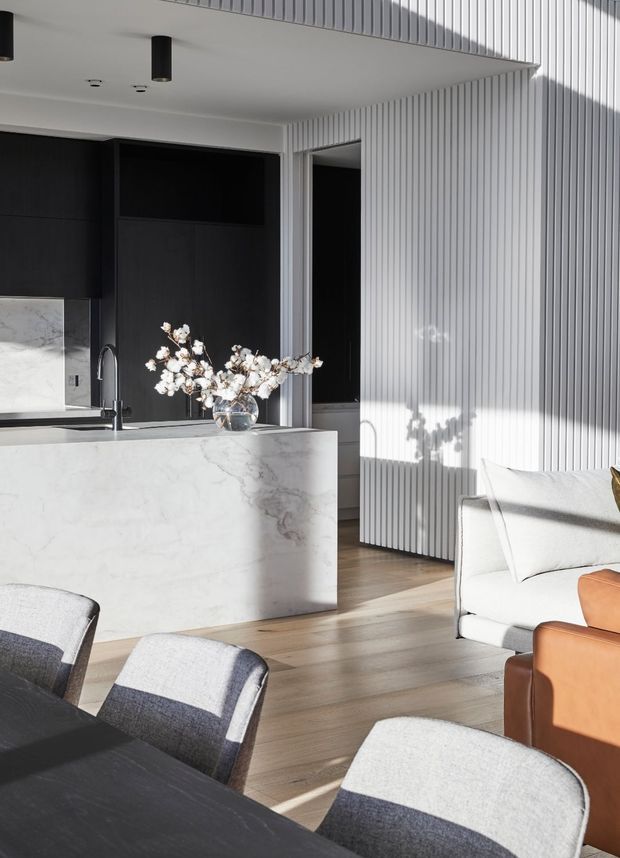 A bright, refined open-plan interior combining a contemporary kitchen, dining area and living room. The kitchen features a large white marble island with integrated sink, dark cabinetry and marble splashback. Vertical white wall panelling, pale timber flooring, a white sofa and modern dining chairs create a clean, high-contrast scheme with warm natural light.