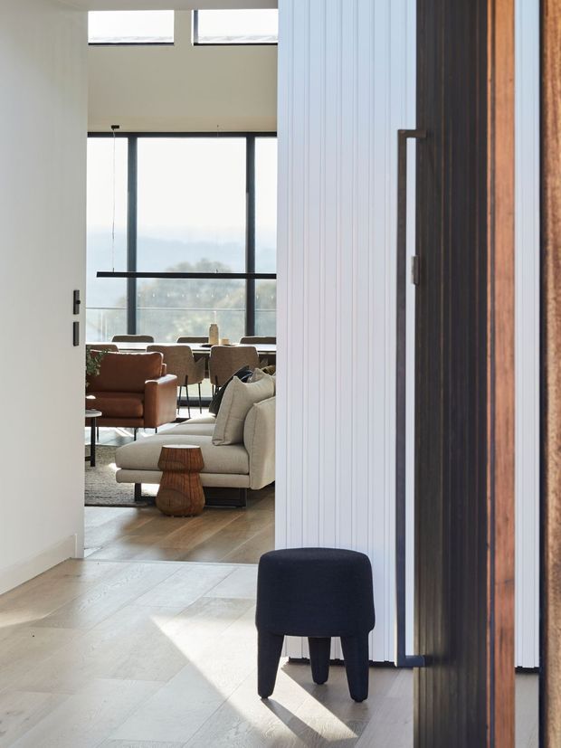Bright contemporary interior view from an entry hallway into an open-plan living and dining area. The foreground features a dark upholstered stool, a wide white door, pale timber flooring, and a vertically ribbed light wall panel beside a dark timber-framed opening. Beyond, a neutral sofa, brown leather armchair, dining table and chairs sit in front of large floor-to-ceiling glazing, with additional high-level windows bringing in abundant daylight.