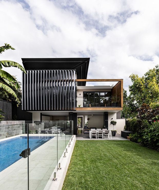 Contemporary two-storey residence viewed from the backyard, featuring a strong black and white facade, deep vertical louvres, timber-lined upper balcony, extensive glazing, and a covered outdoor dining area. A swimming pool with frameless glass safety fencing occupies the foreground, beside a manicured lawn and tropical planting.