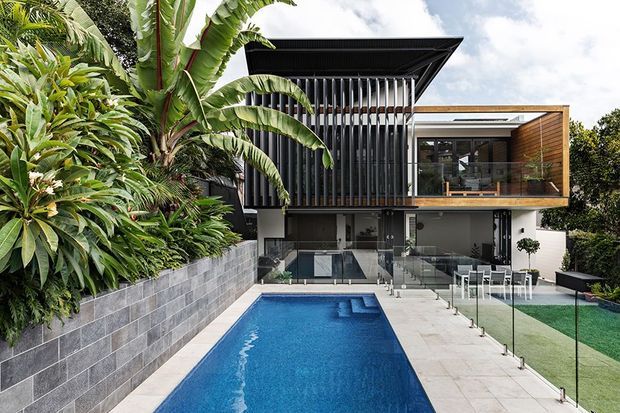 A contemporary multi-level residence viewed from the rear, featuring a long rectangular swimming pool, pale paved terrace, glass balustrades, lush tropical landscaping, dark vertical facade screens, and a projecting timber-clad upper-level balcony. The house has a strongly horizontal composition with a prominent cantilevered upper volume and flat rooflines.