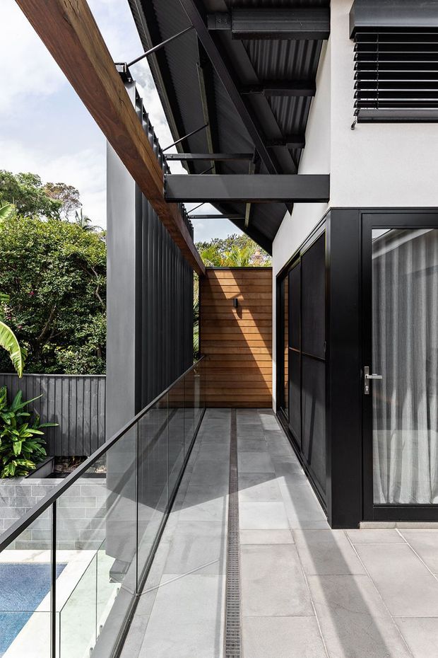 A modern exterior upper-level walkway or balcony beside a house, featuring large grey floor tiles, frameless glass balustrades, black metal-framed doors and window louvers, exposed dark steel roof structure, and a warm timber privacy wall. Lush garden vegetation and a swimming pool are visible below and to the side.