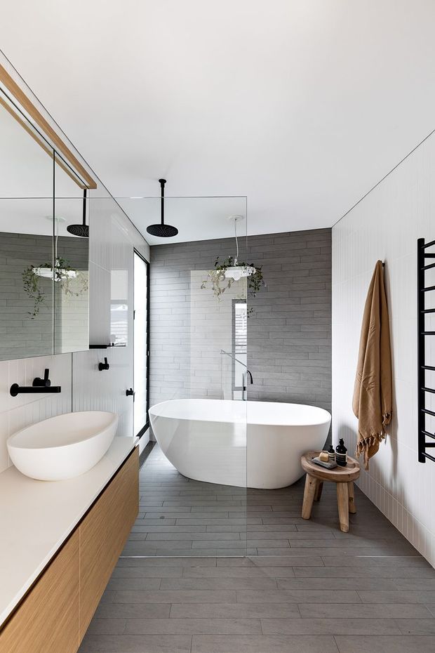 A bright contemporary bathroom featuring a freestanding white bathtub, wall-mounted black fixtures, a large floating timber vanity with vessel basin, frameless glass shower enclosure, grey timber-look floor tiles, grey horizontal wall tiles, and white vertical wall tiles. A black heated towel rail and tan towel are visible on the right wall, with a small wooden stool beside the bath.