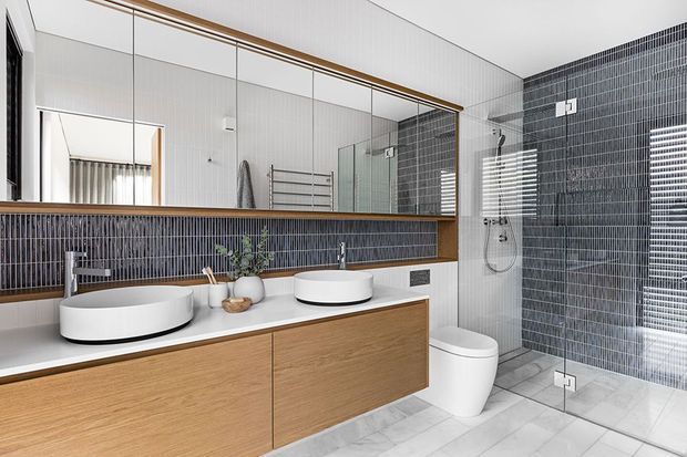 Contemporary bathroom with a long floating timber vanity, two white vessel basins, large mirrored wall cabinetry, and a walk-in glass shower. The room features dark vertically lined wall tiles, pale floor tiles, and restrained black, white, and timber finishes.
