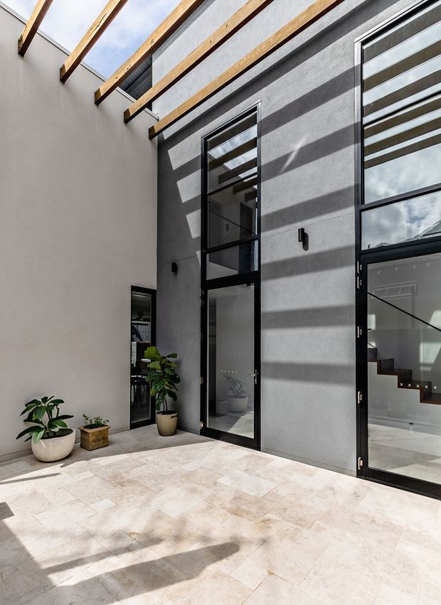 A bright contemporary courtyard or atrium with pale stone paving, smooth grey rendered walls, tall black-framed glazed doors and windows, and exposed timber beams overhead. Strong sunlight creates dramatic linear shadows across the walls and floor, while potted plants add greenery. Interior spaces and a staircase are visible through the glazing.
