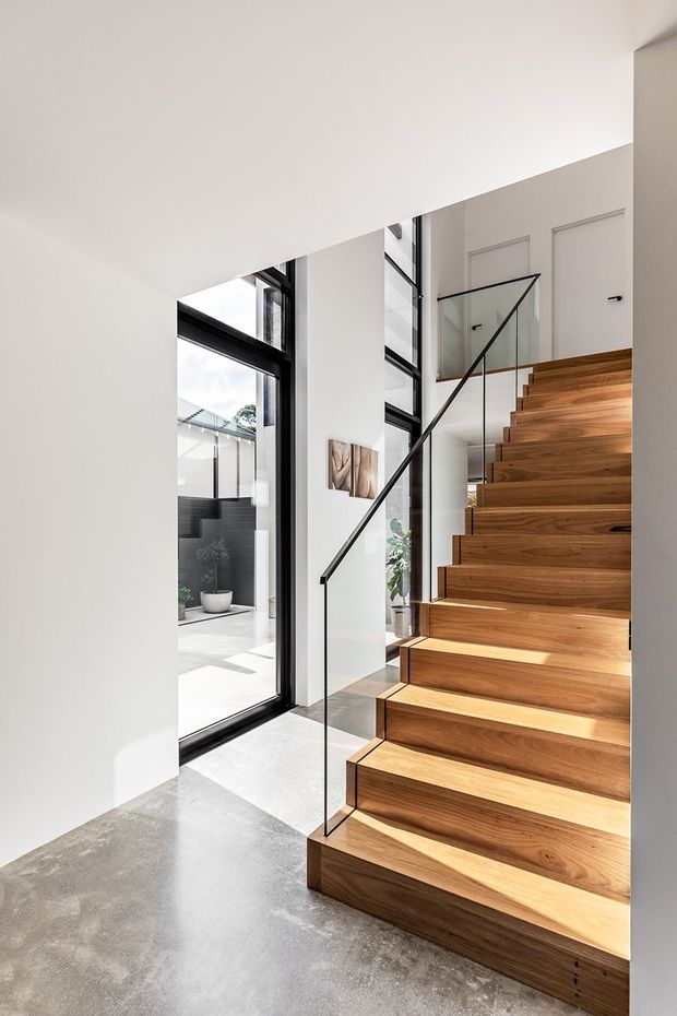 Bright contemporary stairway with warm timber stairs, slim black metal and clear glass balustrade, polished concrete flooring, white walls, and large black-framed floor-to-ceiling glazing opening toward a courtyard. The stair rises to a small upper landing with white panelled doors.