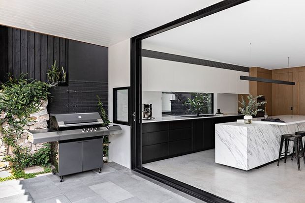 A contemporary indoor-outdoor kitchen viewed from a covered patio or terrace. The foreground features a built-in-style freestanding barbecue beside a dark vertical timber feature wall, stone landscaping and planting. Through large black-framed glazed doors is a bright kitchen with matte black cabinetry, a white marble-look island with waterfall ends, timber cabinetry, integrated appliances and black bar stools. The space has a clean, refined monochromatic palette with warm timber accents.