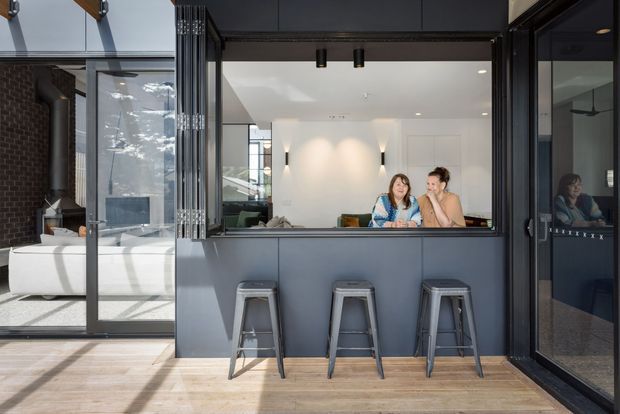 A contemporary indoor-outdoor entertaining area featuring a dark-framed servery or bar opening with a built-in counter and three metal stools. Through the opening is a bright living area with white walls, recessed lighting, a sofa, and two people seated at the counter. Full-height glazing and sliding doors connect the space to an adjacent patio or terrace, while timber flooring, dark cabinetry or wall panels, and exposed brick add material contrast.