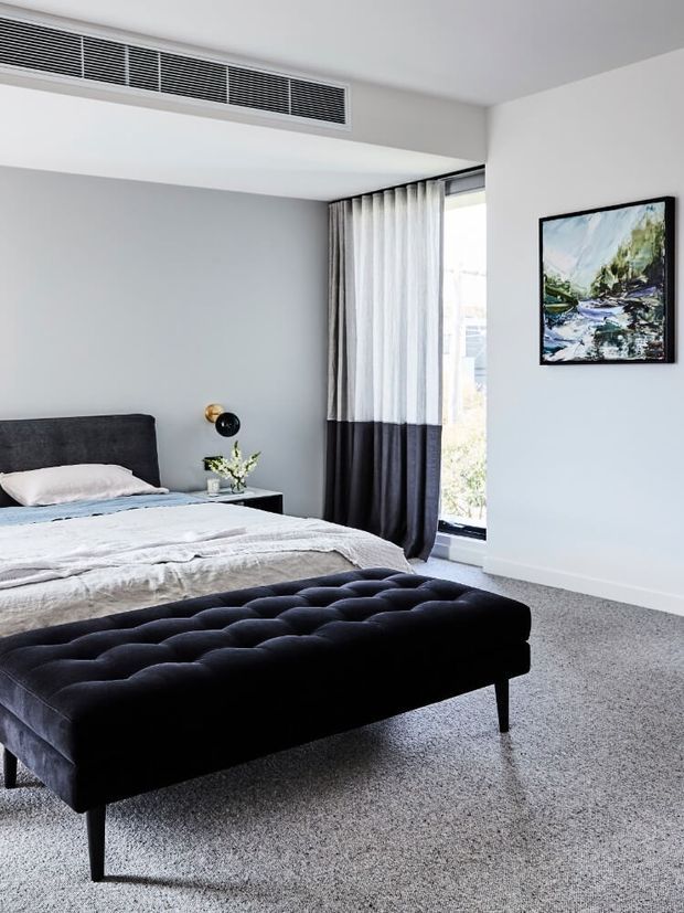 A bright, contemporary bedroom with pale grey walls, grey carpet, a dark upholstered bed and tufted bench, floor-to-ceiling curtains, and a large vertical window. A small bedside table with a wall-mounted light and artwork complete the refined, restrained interior.