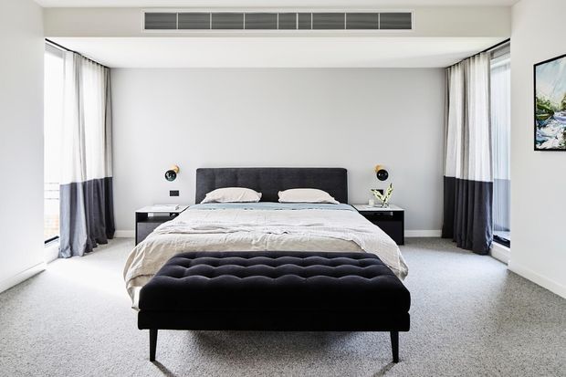 Bright, spacious contemporary bedroom with a centered upholstered bed, dark bedside tables and wall-mounted lamps, a tufted black bench at the foot of the bed, pale carpet, white walls, and full-height windows dressed with sheer and dark curtains. A linear ceiling air-conditioning vent and framed artwork complete the clean, refined interior.