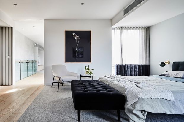 Bright contemporary bedroom with a low upholstered bed, textured light bedding, a large black tufted bench, pale carpet, timber flooring, white walls, sheer curtains with dark lower panels, and a sculptural lounge chair. A glass balustrade and hallway are visible to the left, with minimalist wall art and bedside lighting.