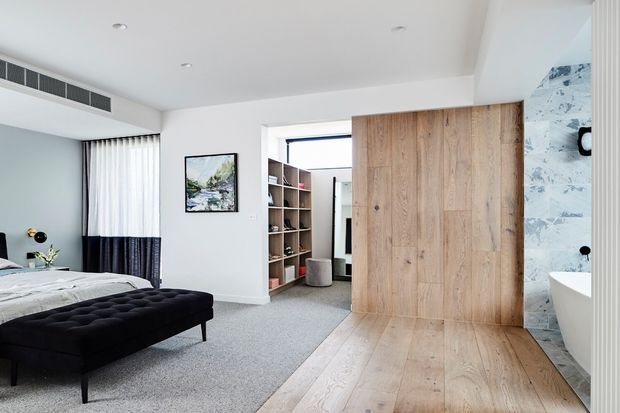 Contemporary bedroom with a large black tufted bench at the foot of the bed, pale carpet, and a wide natural timber sliding door opening to a walk-in wardrobe. A marble-look tiled bathroom with a freestanding white bath is visible at the right, while sheer and dark curtains, recessed lighting, and streamlined white walls create a clean modern interior.