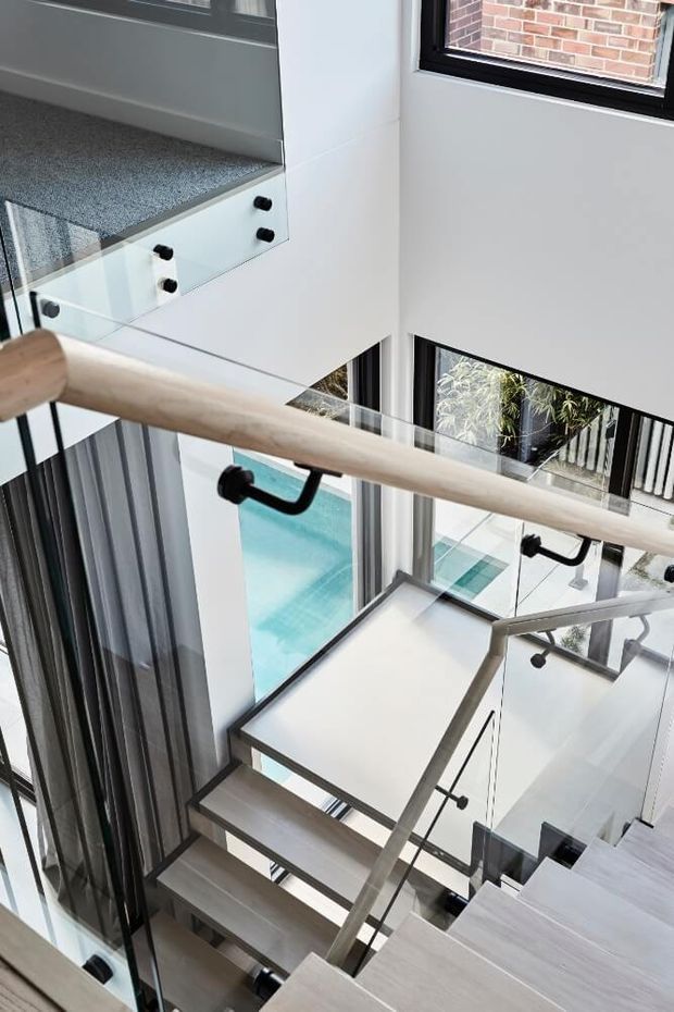 A bright contemporary stairway viewed from above, featuring pale timber open-tread stairs, clear glass balustrades with black fittings, and a light timber handrail. The stairwell opens onto a double-height interior with extensive black-framed glazing, white walls, and views of a turquoise swimming pool and planted courtyard below. A carpeted landing and built-in white joinery are visible at the upper left.