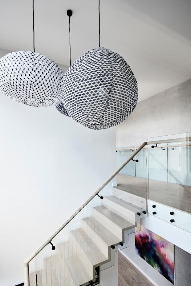 Bright contemporary stairway interior featuring a floating timber staircase with clear glass balustrades and slim metal handrails. Large suspended black-and-white patterned pendant lights dominate the upper frame, while a colourful artwork is visible below the stairs. The space has white walls, light timber flooring and a clean minimalist aesthetic.