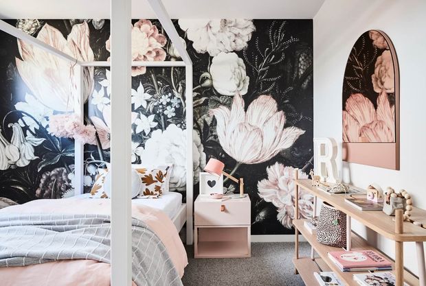 A bright, playful bedroom featuring a four-poster bed, soft blush and grey bedding, and dramatic oversized floral wallpaper in black, white, and pink. Pale timber shelving and a compact bedside table provide storage and display, with arched floral wall panels adding a distinctive decorative feature.