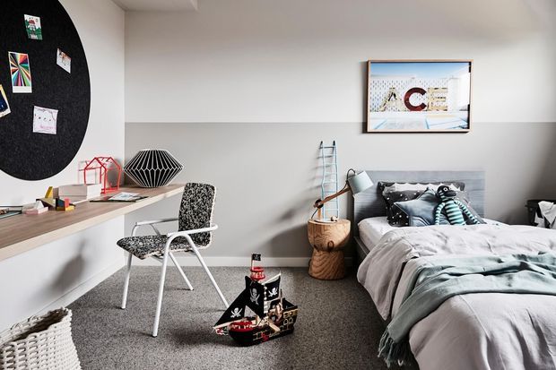 A bright, compact children's bedroom with a single bed, soft grey bedding, a patterned chair at a wall-mounted desk, playful toys, wall art, a large black pinboard, and eclectic decorative objects. The room has pale two-tone walls, grey carpet, and a clean contemporary layout with playful accents.