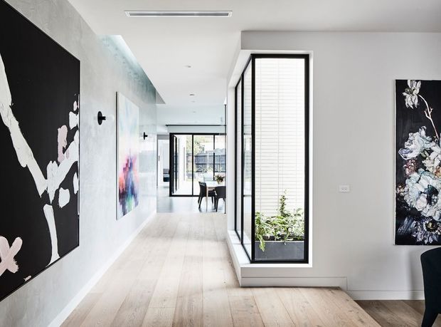 Bright contemporary residential hallway with pale timber flooring, white walls, black-framed floor-to-ceiling glazing, an integrated planter window, recessed ceiling lighting, and large abstract artwork. A dining area is visible beyond the corridor.