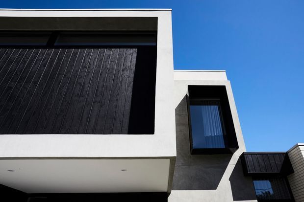 Close-up exterior view of a modern multi-level building with crisp white rendered geometric volumes, dark vertical cladding, projecting black-framed windows, and a flat roofline against a clear blue sky.