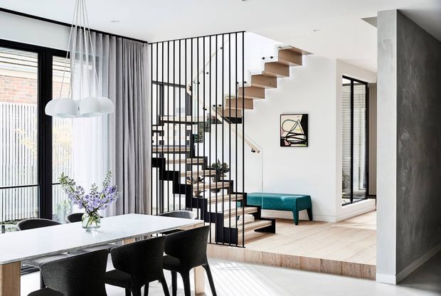 Bright contemporary open-plan interior featuring a dining area with a pale stone table and black upholstered chairs, a sculptural cluster pendant light, and a timber-and-black metal staircase with vertical balustrade screening. A raised living area includes a compact teal bench, while full-height black-framed glazing, sheer curtains, pale timber flooring, white walls, and a dark textured feature wall define the space.