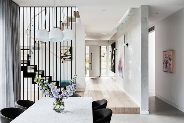 Bright contemporary open-plan entry and dining space with a sculptural black open-riser staircase enclosed by vertical metal balusters, a cluster of white globe pendant lights, pale timber flooring, and a white stone dining table with black chairs. A long hallway extends toward the entry door, with large-scale artwork and textured light walls.