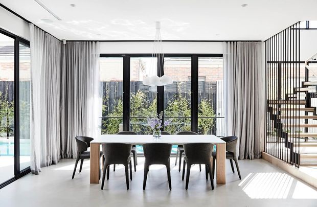 Bright contemporary dining area with a long light-toned dining table and dark upholstered chairs, positioned beside expansive black-framed glazing and full-height grey curtains. A sculptural pendant light hangs above, while a black vertical-screen stair balustrade and open staircase are visible to the right. The pale floor, white ceiling, abundant daylight, and glimpses of greenery and a pool create an airy, refined interior.