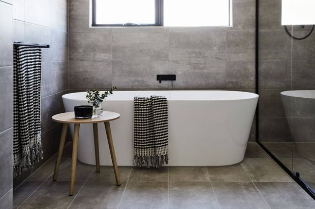 Modern bathroom featuring a freestanding white bathtub against large grey stone-look tiled walls and floor. A black wall-mounted tap, frameless glass shower screen, high horizontal window, and timber-legged side table with monochrome textiles complete the minimalist setting.