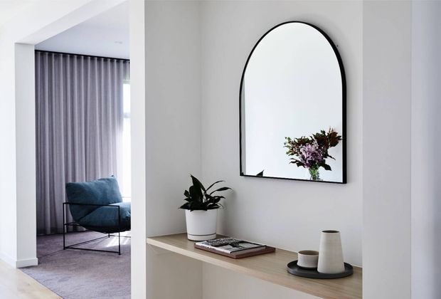 A refined, minimalist entryway with a pale built-in console, arched black-framed mirror, potted plant, books, and ceramic vessels. An adjacent sitting area is visible through the opening, featuring a blue lounge chair, floor-to-ceiling curtains, and carpeted flooring. The palette is predominantly white, light grey, and light timber with restrained black accents.