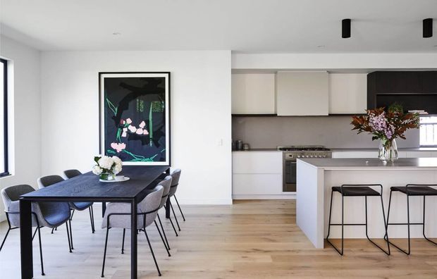 A bright open-plan dining and kitchen interior. A dark rectangular dining table with upholstered chairs occupies the foreground, while a white island with black bar stools, pale cabinetry, stainless-steel appliances, and a floral arrangement defines the kitchen behind it. Light timber flooring, white walls, black-framed artwork, and minimal black ceiling lights create a clean contemporary look.