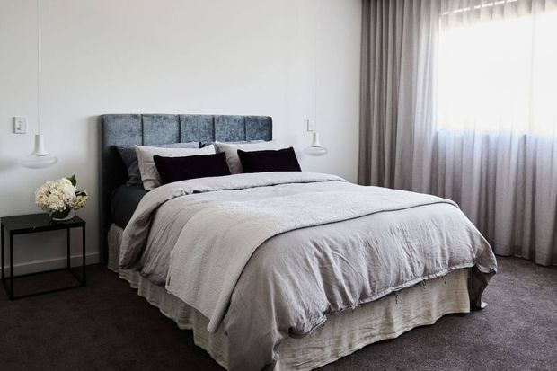 A softly lit contemporary bedroom featuring a neatly made bed with a tall upholstered grey headboard, layered white and grey bedding, dark accent cushions, bedside tables with pendant lights, and full-height sheer and blackout curtains. Dark carpet and neutral finishes create a calm, refined atmosphere.