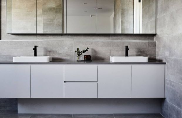 Contemporary double-vanity bathroom with a long floating white vanity, two white vessel basins, matte black wall-mounted taps, a dark benchtop, large grey tiled walls and floor, and a wide mirrored cabinet above.