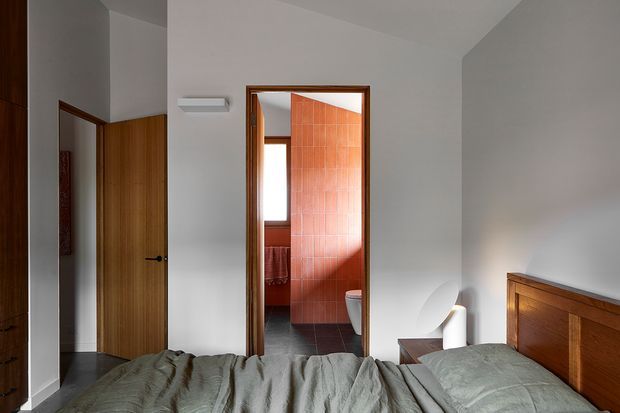 A calm, contemporary bedroom viewed toward an open bathroom doorway. The room has white walls, dark flooring, a timber door and built-in timber headboard, with muted green-grey bedding. The bathroom beyond features vivid terracotta-red tiled walls, dark floor tiles, a toilet, and a small window. Natural light enters from the bathroom and brightens the otherwise restrained interior.