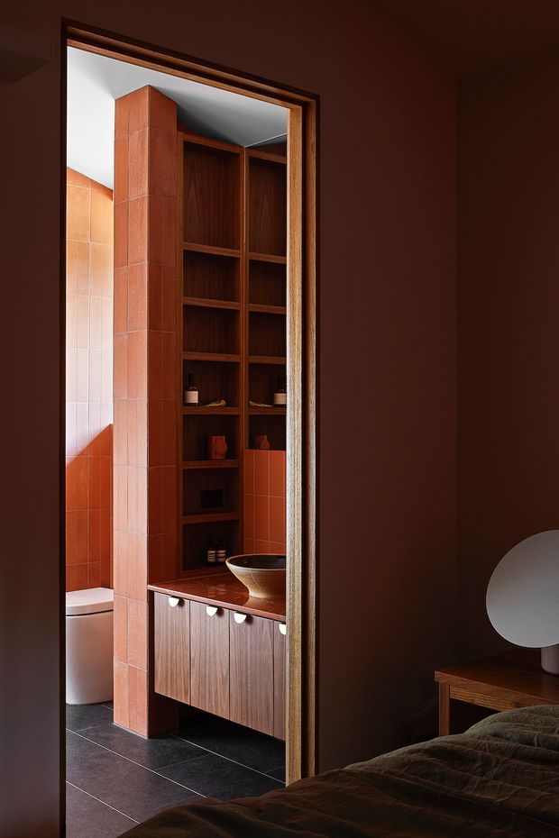 A moody interior view from a bedroom through an open doorway into a compact bathroom. The bathroom features terracotta-toned tiled walls, dark tiled flooring, a warm timber vanity with integrated basin and open timber shelving, while the foreground bedroom has deep brown walls, a timber side table, sculptural lamp, and dark bedding. The doorway and timber detailing create a strong framed view between the two spaces.