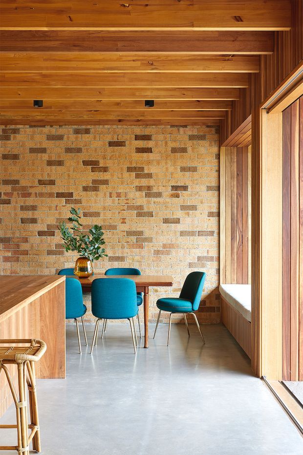 A warm, compact dining area with a timber-lined ceiling and built-in timber joinery, exposed brick feature wall, polished concrete floor, and large timber-framed glazing along the right side. A small wooden dining table is surrounded by vivid teal upholstered chairs, with a partial kitchen or cabinetry zone in the foreground. The space has strong natural light, exposed structural detailing, and a restrained mid-century-inspired palette.