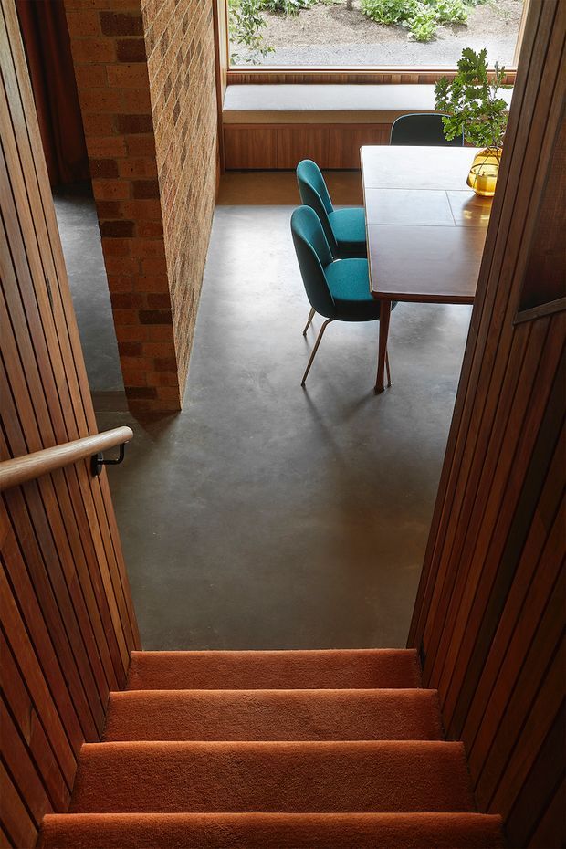 View down a carpeted staircase into a compact dining area. The space features warm timber vertical wall panelling, exposed red brick, a polished concrete floor, a mid-century-style wooden dining table with teal upholstered chairs, and a built-in window seat beneath a large garden-facing window. A handrail and stair trim frame the foreground.