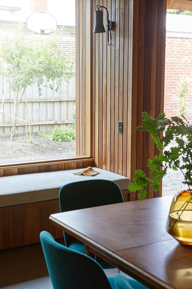 A warm, compact dining nook beside a large window, featuring vertical timber wall panelling, a built-in upholstered window seat, a dark wood dining table, teal upholstered chairs, and a wall-mounted black metal pendant or sconce. The space looks into a planted courtyard and receives abundant natural light. A sculptural amber glass table lamp adds a warm accent.