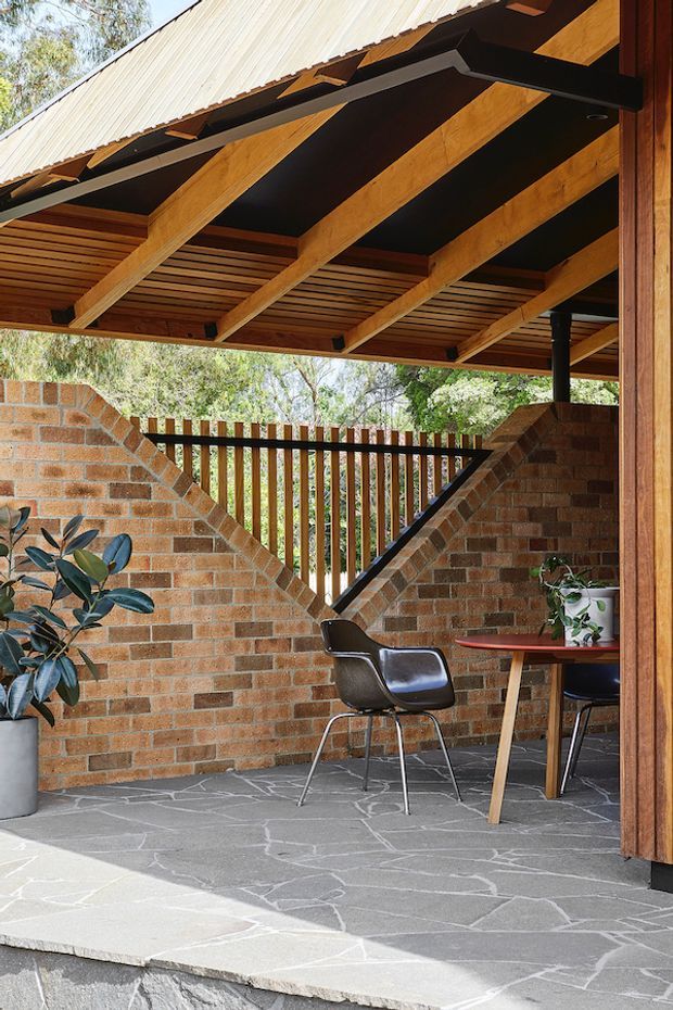 A covered outdoor patio or terrace with a timber-framed pitched roof, exposed beams and timber soffit, warm brick walls, dark slate-like irregular stone paving, and a timber slatted balustrade. The space contains a small dining table and chairs, with potted greenery. The architecture combines robust masonry with warm natural timber in a sheltered garden setting.