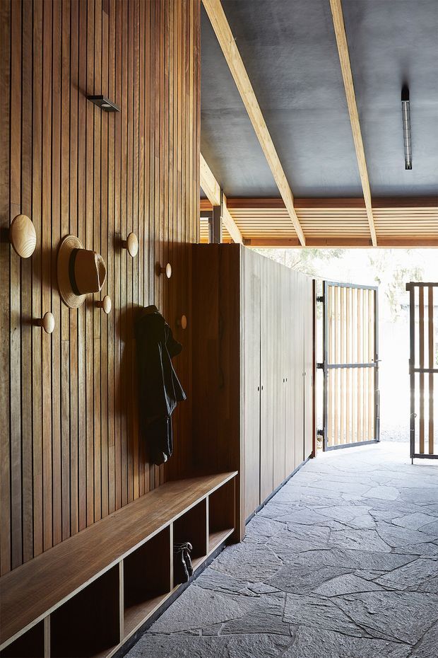 A warm, timber-lined entryway or mudroom with a full-length built-in bench, open cubby storage, coat hooks, and tall concealed cabinetry. Vertical timber battens cover the feature wall and continue across the ceiling, contrasting with exposed pale timber beams and a dark grey ceiling. The space opens to a gated exterior, with irregular natural stone paving underfoot and abundant daylight.