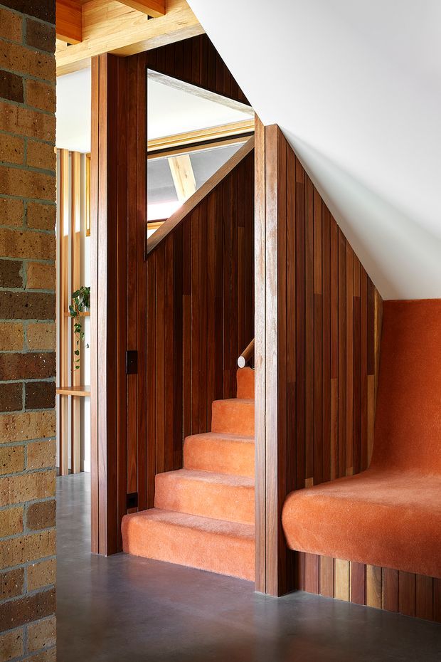 Interior view of a compact stairway enclosed by rich vertical timber panelling, with warm orange carpeted stairs and an integrated orange upholstered bench or seating area beneath a sloping ceiling. Exposed brickwork frames the left side, while timber beams, skylight-like glazing and glimpses of an adjacent space add architectural interest. The palette is warm, earthy and strongly material-focused.