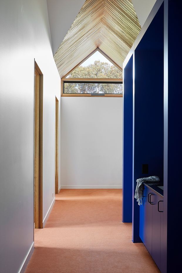 A narrow, contemporary residential hallway or landing with pale carpet, white walls, warm timber door frames, and vivid deep-blue built-in cabinetry along the right side. The corridor terminates in a distinctive high triangular gable window beneath a timber-lined vaulted ceiling, bringing in natural light and views of trees. Minimal fixed detailing and strong blue-and-white contrast define the space.