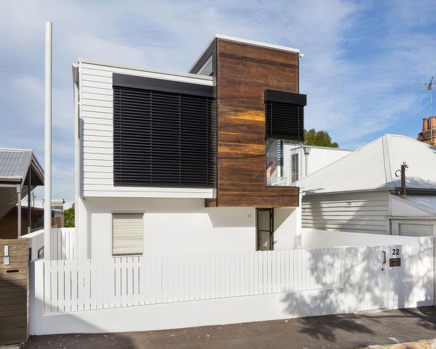Street-facing view of a contemporary multi-level residence in an established suburban setting. The facade combines white horizontal cladding and rendered walls with a prominent vertical feature clad in dark timber boards. Large windows are screened with black horizontal louvres, and the property is enclosed by a white vertical-slat fence and gate. Adjacent pitched-roof homes are visible on either side.