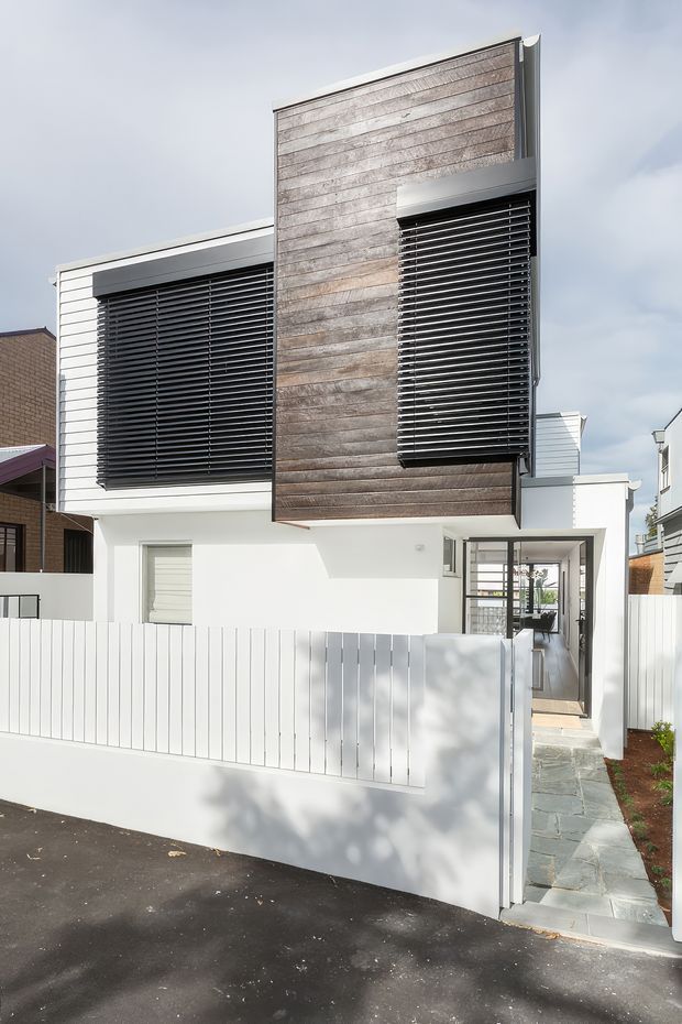 Street-facing view of a contemporary multi-storey townhouse with a crisp white rendered facade, prominent weathered timber-clad upper volume, and large horizontal dark louvres providing solar shading. A narrow paved entry path leads through a white vertical-slat fence to the recessed front door. The composition highlights the building envelope and facade detailing.