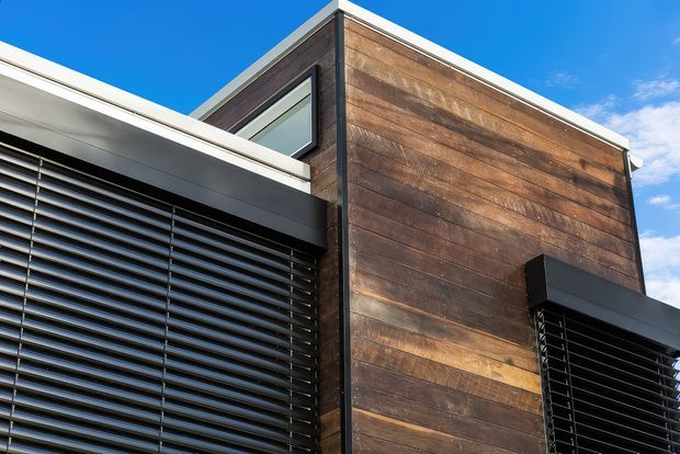 Close-up exterior architectural photograph of a contemporary building corner. The facade combines broad horizontal dark timber cladding with crisp white roofline and trim, dark metal-framed glazing, and large black horizontal louvres providing solar shading. A vivid blue sky is visible above.