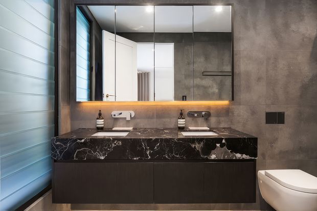 A contemporary bathroom featuring a wall-mounted dark timber vanity with a dramatic black marble benchtop, twin basins and wall-mounted taps. Large mirrored cabinets span the vanity, with warm integrated underlighting. Large grey concrete-look wall tiles, a louvred window, and a white toilet create a refined modern industrial aesthetic.